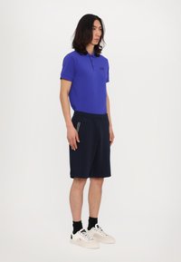 Polo di colore blu royal con colletto e logo sul petto, abbinato a pantaloni corti blu navy e sneakers bianche, caratterizzato da una texture liscia.