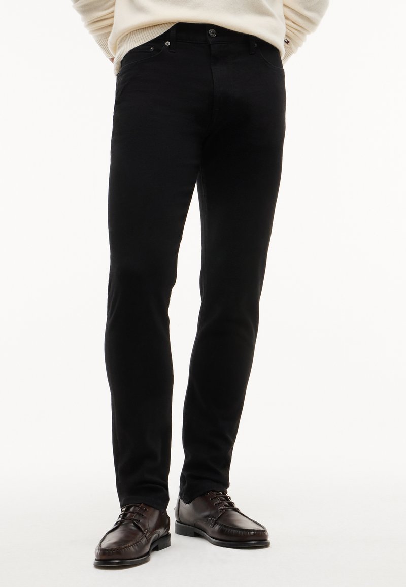 Άνδρας με μαύρο τζιν slim-fit και σκούρα καφέ δερμάτινα δετά παπούτσια, μπροστά σε λευκό φόντο.