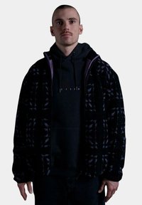 Chaqueta de forro polar negra con acentos morados, que presenta un patrón de cuadros. Llevada sobre una sudadera negra con capucha, con un ajuste relajado y puños de canalé.