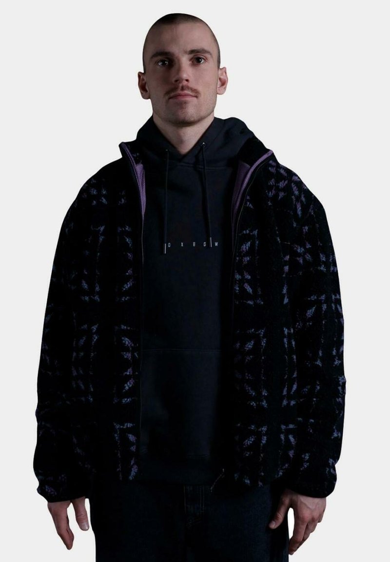 Chaqueta de forro polar negra con acentos morados, que presenta un patrón de cuadros. Llevada sobre una sudadera negra con capucha, con un ajuste relajado y puños de canalé.