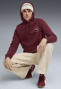 Burgundy hættetrøje med forside-logo, cremet hue med sorte striber, beige bukser og maroon sneakers, der har et klassisk atletisk design.