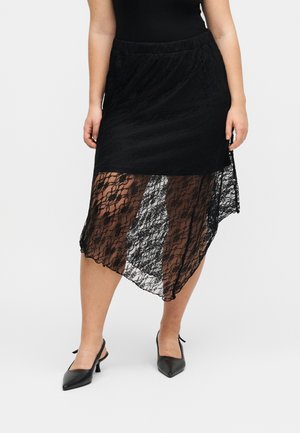 Femme portant une jupe en dentelle noire asymétrique avec une sous-couche solide, associée à des talons slingback noirs, debout sur un fond uni.