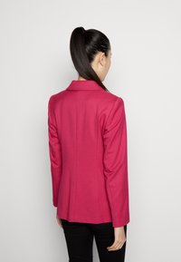 Blazer rosa aderente con colletto a punte, maniche lunghe e dettagli cuciti, realizzato in tessuto liscio. Indossato con pantaloni neri.