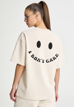 Frau mit langen Haaren im Pferdeschwanz, die eine Brille trägt und ein beigefarbenes Hemd mit einem schwarzen Smiley-Gesicht und dem Text "I DON'T CARE" auf dem Rücken.