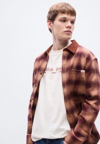 Κόκκινο και καφέ καρό πουκάμισο από φανέλα με καφέ γιακά, φορεμένο πάνω από ένα λευκό μπλουζάκι με κόκκινες επιγραφές. Μπλε τζιν και διακριτικές λεπτομέρειες στις τσέπες εμφανείς.