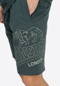 Mørkegrønne bomuldsshorts med løs pasform. Har et stort hvidt løvegrafik og teksten "Lonsdale London" på venstre ben.