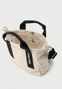 Μπεζ τσάντα από καμβά με μαύρες λεπτομέρειες, διαθέτει κοντές λαβές και ρυθμιζόμενο ιμάντα. Στο εσωτερικό φαίνεται ετικέτα με το λογότυπο "Calvin Klein".
