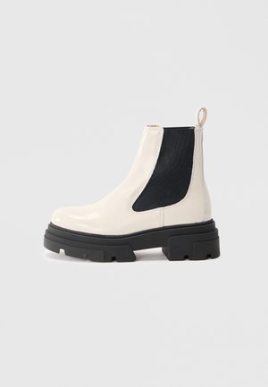 Bottines Chelsea en cuir crème avec semelle noire épaisse et panneau latéral élastique noir sur fond blanc uni.