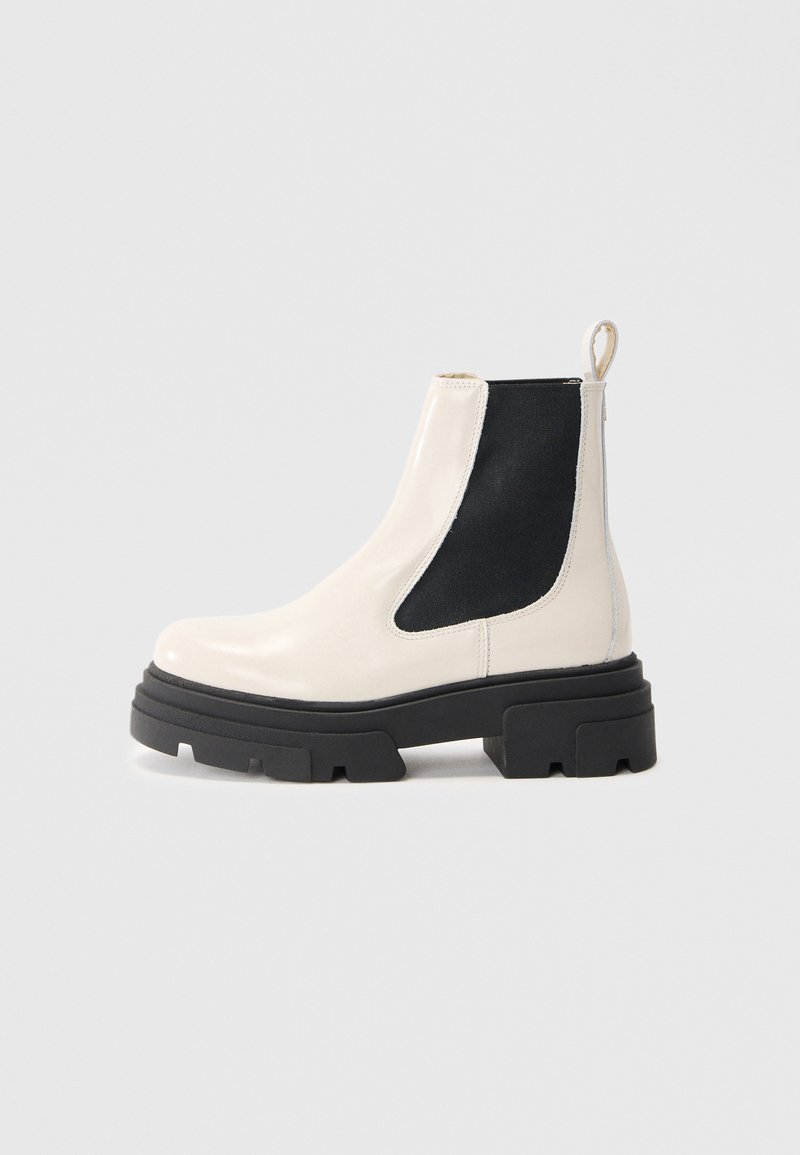 Bottines Chelsea en cuir crème avec semelle noire épaisse et panneau latéral élastique noir sur fond blanc uni.
