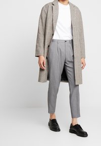 Lange geruite jas in beige, grijs en rode patronen, gedragen over een wit T-shirt. Grijze op maat gemaakte broek en zwarte veterschoenen maken de outfit compleet.