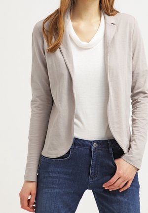 Kvinde iført beige åben blazer over hvid top med blå jeans, hånd hvilende nær lommen foran på hoften.