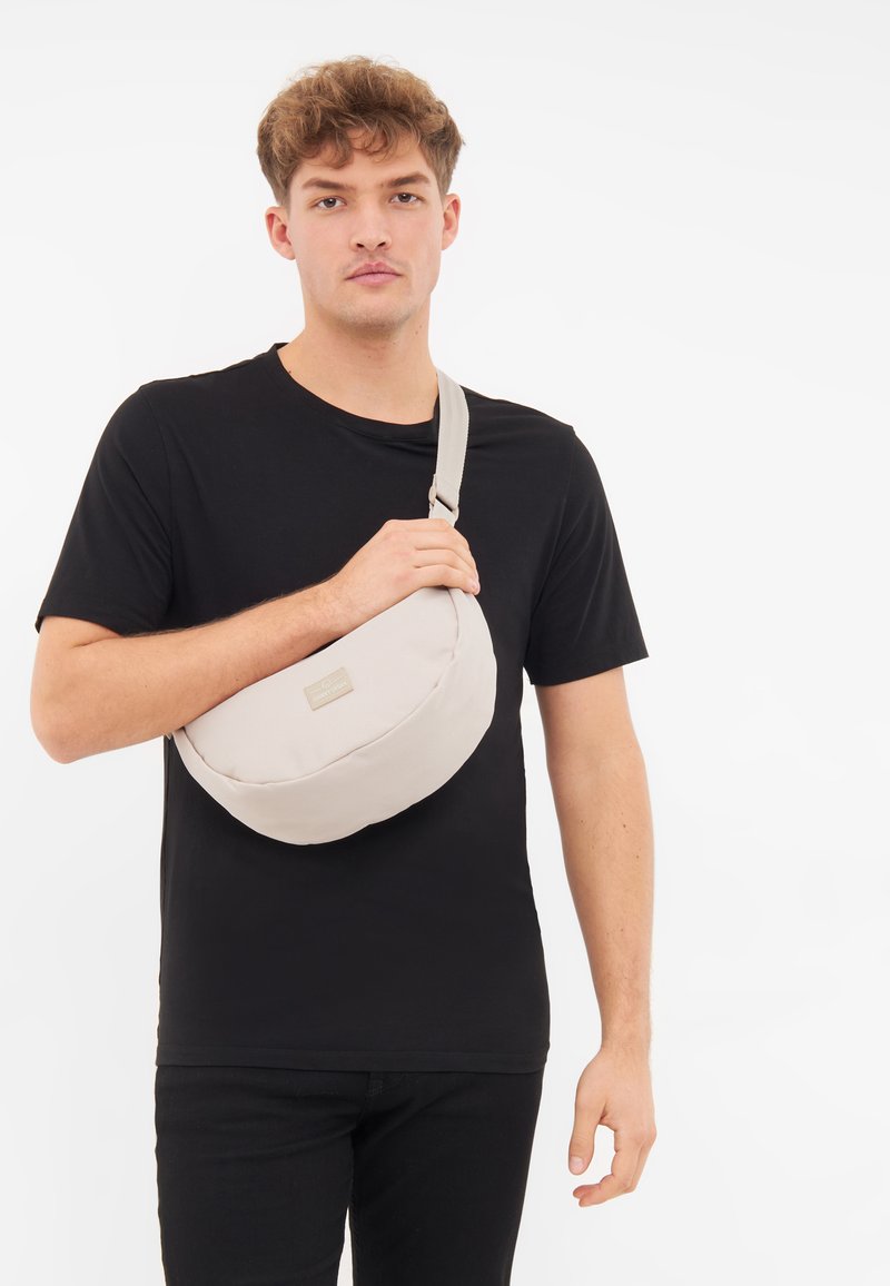Borsa a tracolla beige con finitura in tessuto morbido, design curvo, tracolla regolabile e un piccolo logo impresso sul davanti.