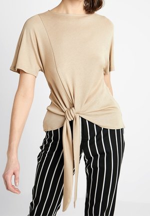 Haut beige à manches courtes avec détail de nœud à l'avant, porté avec un pantalon noir à rayures verticales blanches sur une personne debout.