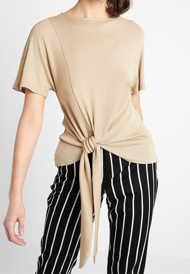 Haut beige à manches courtes avec détail de nœud à l'avant, porté avec un pantalon noir à rayures verticales blanches sur une personne debout.