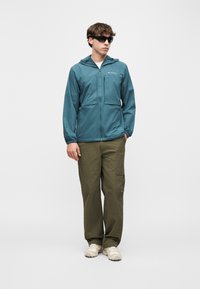 Νέος άνδρας με μπλε φλις μπουφάν με φερμουάρ και κουκούλα, χακί παντελόνι cargo, λευκά αθλητικά παπούτσια και μαύρα γυαλιά ηλίου, στέκεται μπροστά σε αδιάφορο φόντο.