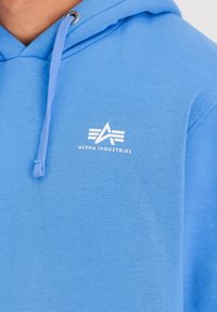 Svetlo modra kapuca iz mehkega materiala z belim logotipom spredaj. Ima vrvico na kapuci in teksturirano površino.