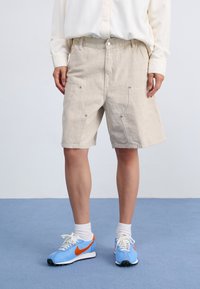 Beige, knälånga shorts gjorda av strukturerad tyg med två framfickor med tryckknappdetaljer. Matchas med blåa träningsskor.