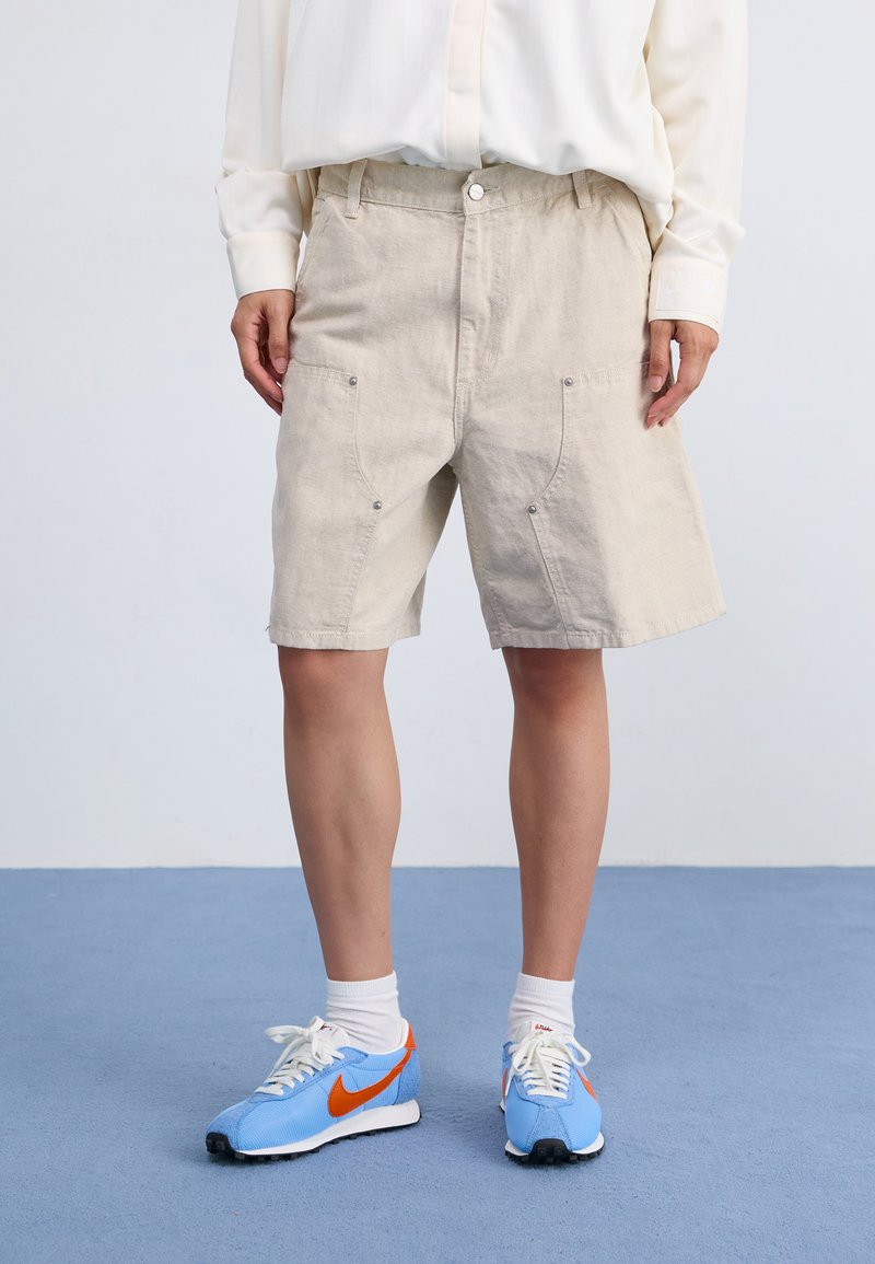 Beige, knälånga shorts gjorda av strukturerad tyg med två framfickor med tryckknappdetaljer. Matchas med blåa träningsskor.