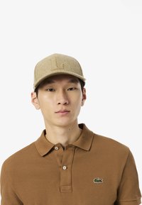 En tanfärgad ullkeps med en svängd skärm och en präglad "L"-logga framtill, tillsammans med en brun pikétröja med en liten grön logga.