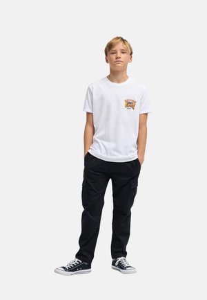 Ragazzo in piedi con le mani in tasca che indossa una t-shirt bianca con grafica, pantaloni cargo neri e sneakers basse nere su sfondo semplice.