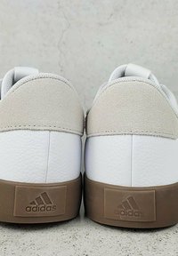 Biele kožené tenisky s béžovou semišovou pätou, textúrovanou gumovou podrážkou a hnedým brandingovým panelom s logom Adidas.