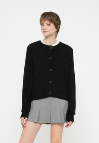 Černý pletený cardigan s kulatým výstřihem, se zapínáním na knoflíky a žebrovanými manžetami. Nesený přes bílou košili a šedou sukni.