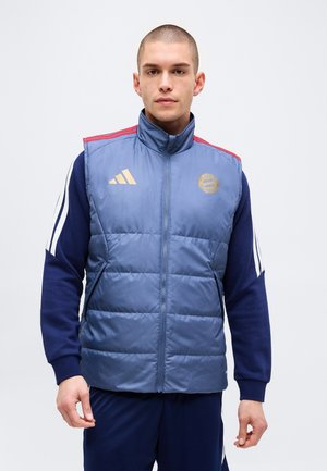 Jonge man in een blauw gewatteerd vest met FC Bayern München en Adidas-logo's, draagt een donkerblauw Adidas trainingspak met witte strepen.
