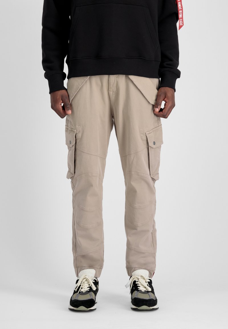 Homme portant un pantalon cargo beige avec plusieurs poches, des baskets noires et blanches, et un sweat noir avec une étiquette rouge sur la manche.