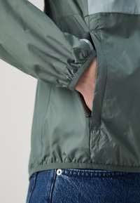 Veste légère verte avec poignets élastiques et une poche latérale. Tissu lisse et brillant avec une coupe ajustée, superposée aux épaules.