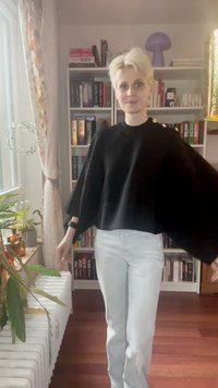 Persoon met kort blond haar die een zwarte trui met gouden knopen en witte broeken draagt, staat in een kamer met boekenkasten, planten en een raam.