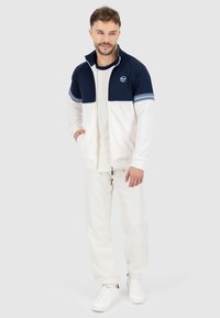 Veste zippée bleu marine et blanche avec un logo, assortie à un pantalon blanc. Présente un design bicolore et des baskets blanches. Tissu doux et décontracté.