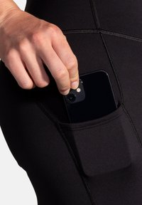 Leggings neri con una tasca laterale, texture liscia e cuciture a contrasto; una mano sta estraendo uno smartphone dalla tasca.