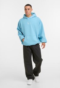 Jauns vīrietis ar īsiem matiem un pūkām, ģērbies gaiši zilā kapučjodā, brīvās melnās džinsās un baltās sporta kurpēs, stāv uz baltās fona.