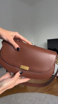 Brun læderhåndtaske med en glat tekstur, buet form og et guldfarvet metalplakette. Har en lang, justerbar rem.