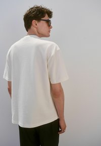 Camiseta color crema con mangas cortas, cuello redondo y un corte holgado. La tela parece suave con una textura lisa.