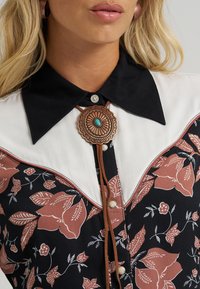 Blusa preta com padrão floral, pala branca, acentos florais castanhos e um colar bolo com uma peça central circular turquesa.