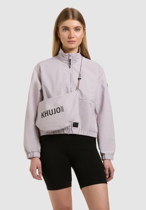 Giovane donna che indossa una giacca con zip di colore viola chiaro e pantaloncini da ciclismo neri, con una borsa a tracolla con la scritta "KHUJO 2001".