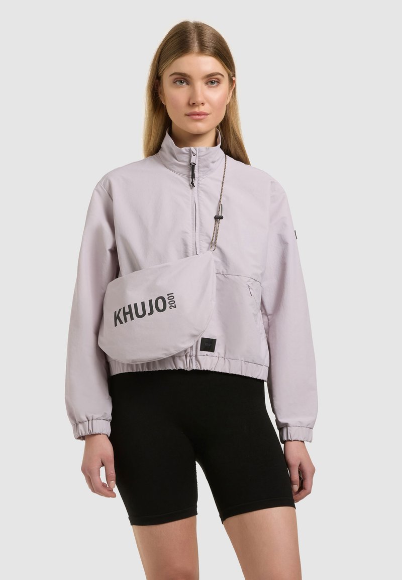 Jeune femme portant une veste zippée violet clair et un short de vélo noir, avec un sac porté en bandoulière arborant le texte "KHUJO 2001".
