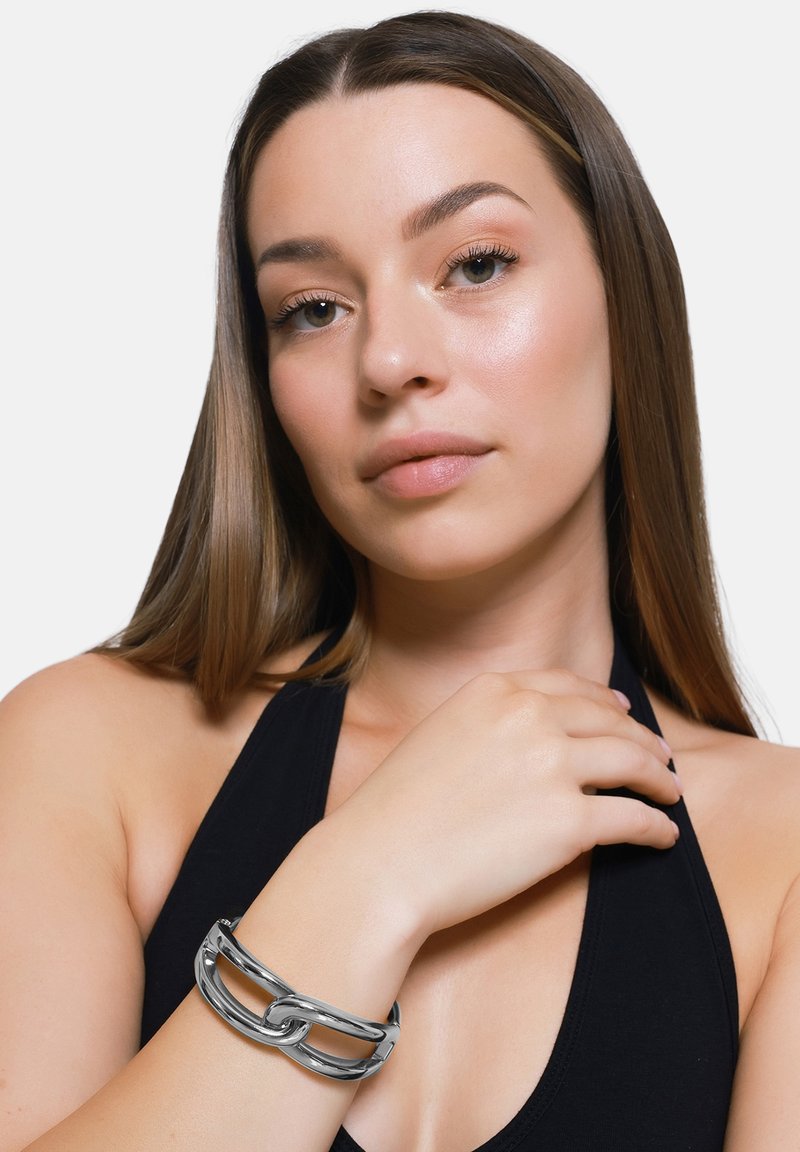 Bracciale in metallo argento con un design a catena intrecciata. Superficie liscia e lucida; indossato su una mano su uno sfondo di abbigliamento nero.