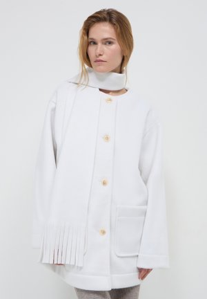 Cappotto di pile bianco con design dritto, grandi bottoni frontali, tasche laterali e un accento frangiato lungo l'orlo. Texture liscia.