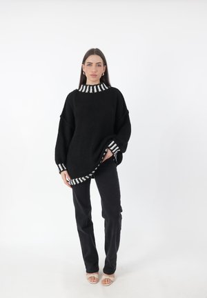 Zwarte gebreide trui met een dikke witte en zwarte gestreepte kraag, oversized ontwerp en contrasterende gestreepte manchetten. Gepaard met zwarte broek en sandalen.