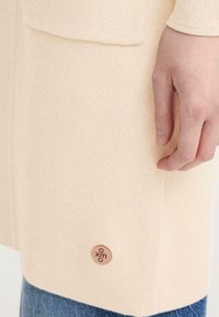 Cardigan beige clair à tissu texturé, avec une poche plaquée et une petite étiquette en cuir circulaire avec un logo embossé sur l'ourlet.
