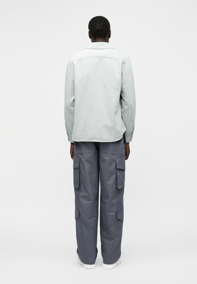 Άνδρας που κοιτάζει προς τα πίσω, φοράει ανοιχτό γκρι μακρυμάνικη μπλούζα και σκούρο γκρι παντελόνι cargo με πολλές τσέπες, στέκεται μπροστά σε απλό φόντο.