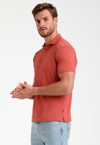 Kortærmet polo shirt i koral, lavet af blødt materiale. Har klassisk krave, knapstolpe og slidser i siden ved kanten.