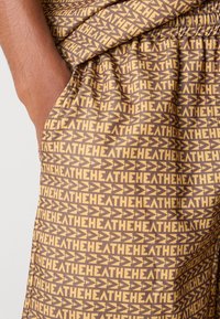Brown shorts with a repeating yellow geometric pattern. Features an elastic waistband and side pockets, made of lightweight fabric.