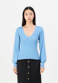 Suéter azul-claro com nervuras, de decote em V profundo e mangas longas, combinado com uma saia preta com botões de latão na parte da frente.