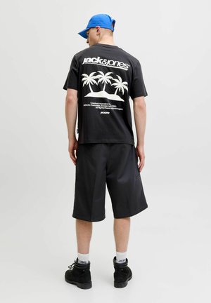 Mand iført sort Jack & Jones t-shirt med hvidt palmetræsgrafik, sorte shorts, sorte støvler, hvide sokker og blå kasket, set bagfra.