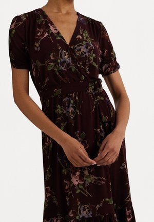 Femme portant une robe portefeuille fleurie foncée à manches courtes avec une ceinture à la taille, mains doucement jointes devant.