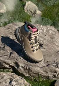 Bota de senderismo beige con textura de malla, detalles en negro y rosa, puntera reforzada y suela de goma, posicionada sobre una superficie rocosa.
