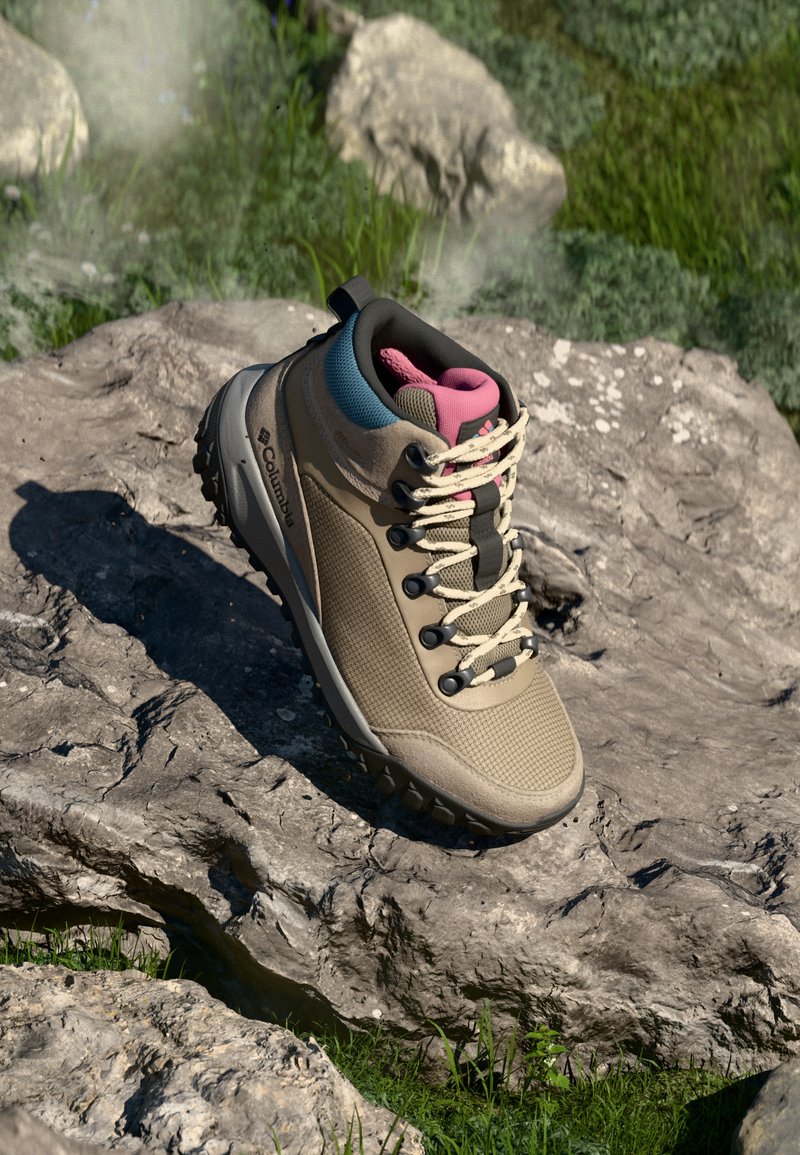 Bota de senderismo beige con textura de malla, detalles en negro y rosa, puntera reforzada y suela de goma, posicionada sobre una superficie rocosa.